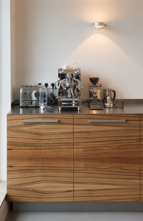 Coffee station on wood-grain kitchen cabinets beneath wall lights.